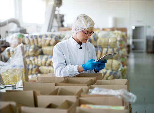 A worker in a white lab coat and blue gloves inspects items on a tablet amid stacks of packaged goods in a warehouse setting.