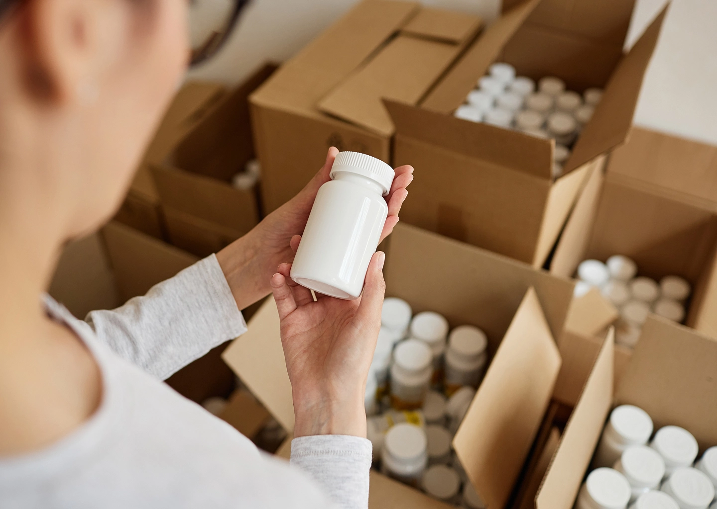 A person holds a white pill bottle in front of several boxes filled with more pill bottles, suggesting an inventory of medications.