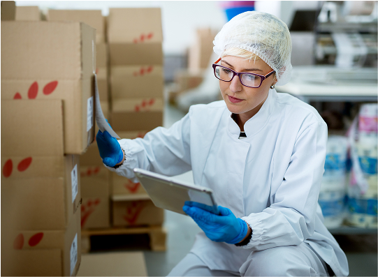 A person in a white lab coat and blue gloves reviews documents on a tablet while seated near stacked cardboard boxes in a workspace.